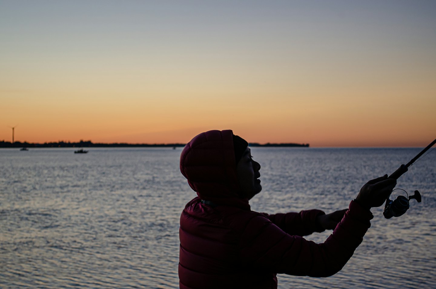 Person fishing at sunrise over calm water with silhouetted boats.